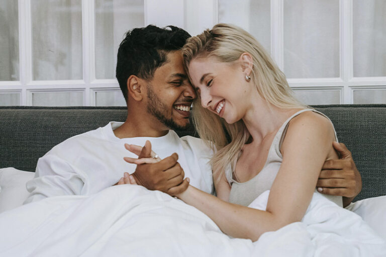 Couple holding hands and laughing together in bed, represents how sex and intimacy therapy can help you and your partner reconnect.